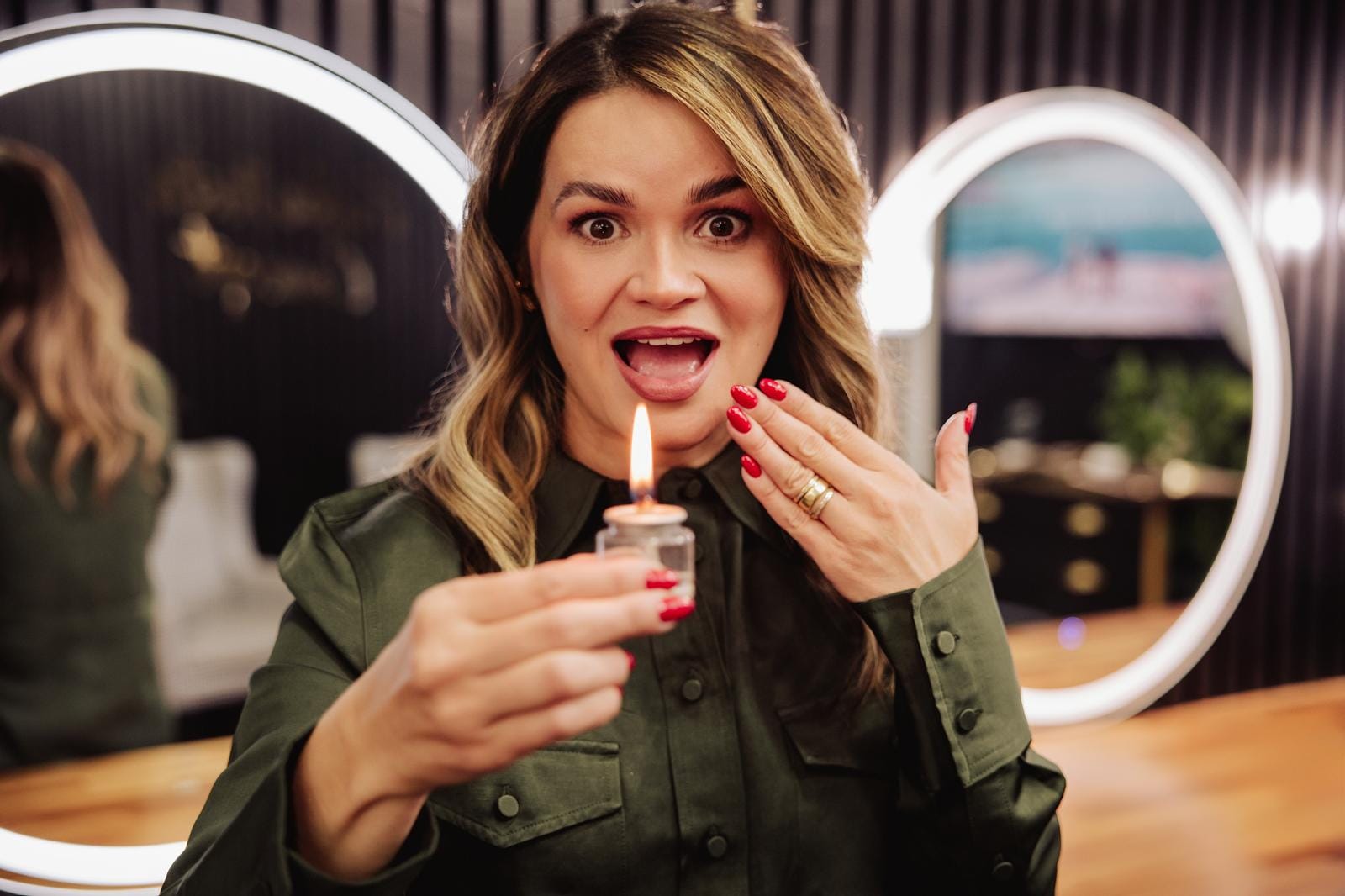 Erika Mota rindo, segurando uma vela acesa em frente aos espelhos iluminados do salão