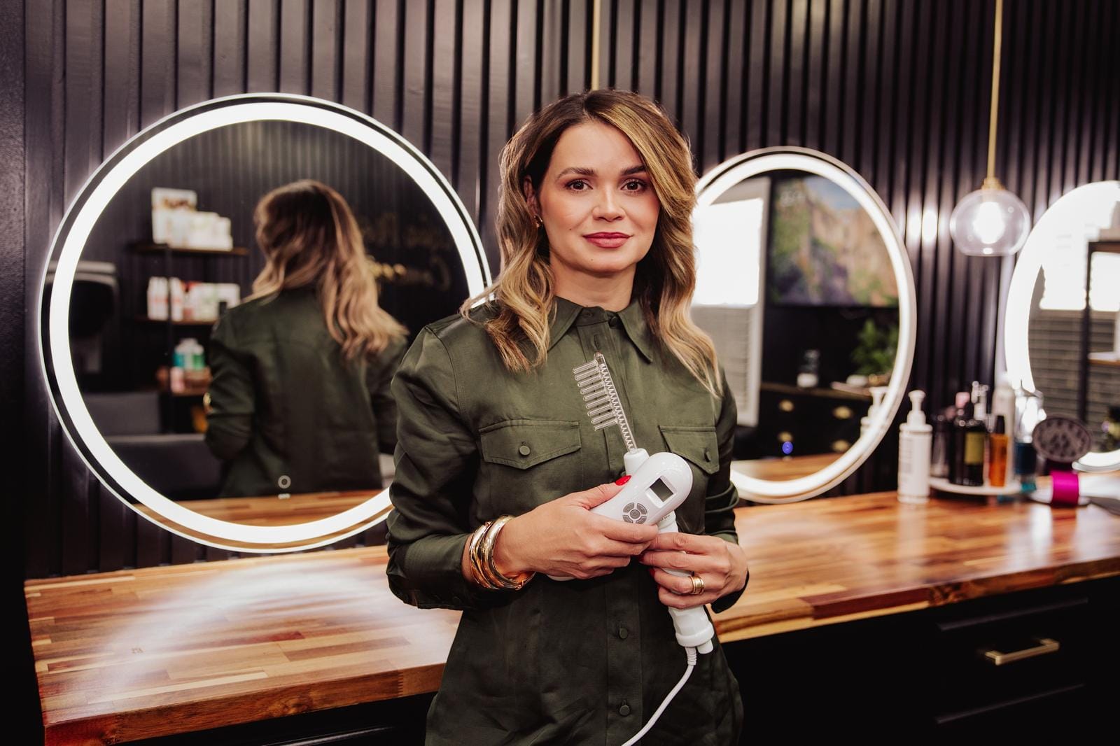 Erika Mota na bancada de trabalho do salão, com secador na mão e espelhos redondos iluminados