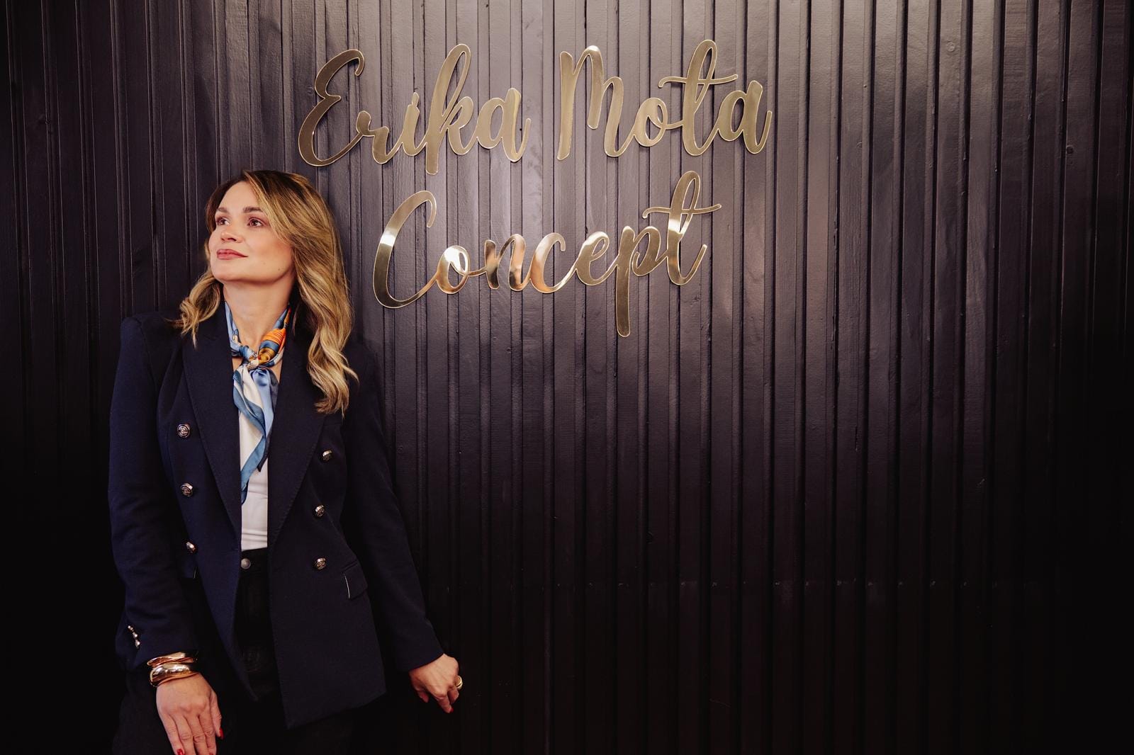 Erika Mota de blazer azul-marinho em pé diante do letreiro dourado Erika Mota Concept