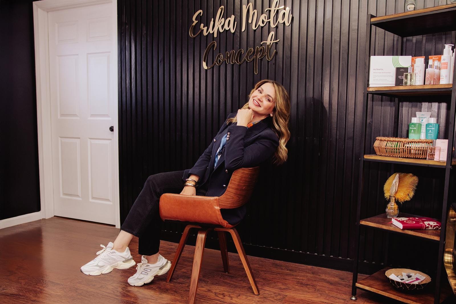 Erika Mota sentada numa cadeira de couro em frente à parede ripada preta com o letreiro dourado Erika Mota Concept, ao lado da estante de produtos