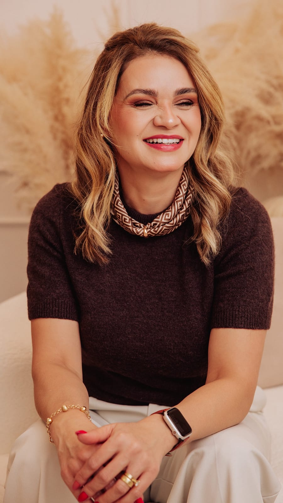 Erika Mota sorrindo, sentada, de blusa marrom e colar dourado