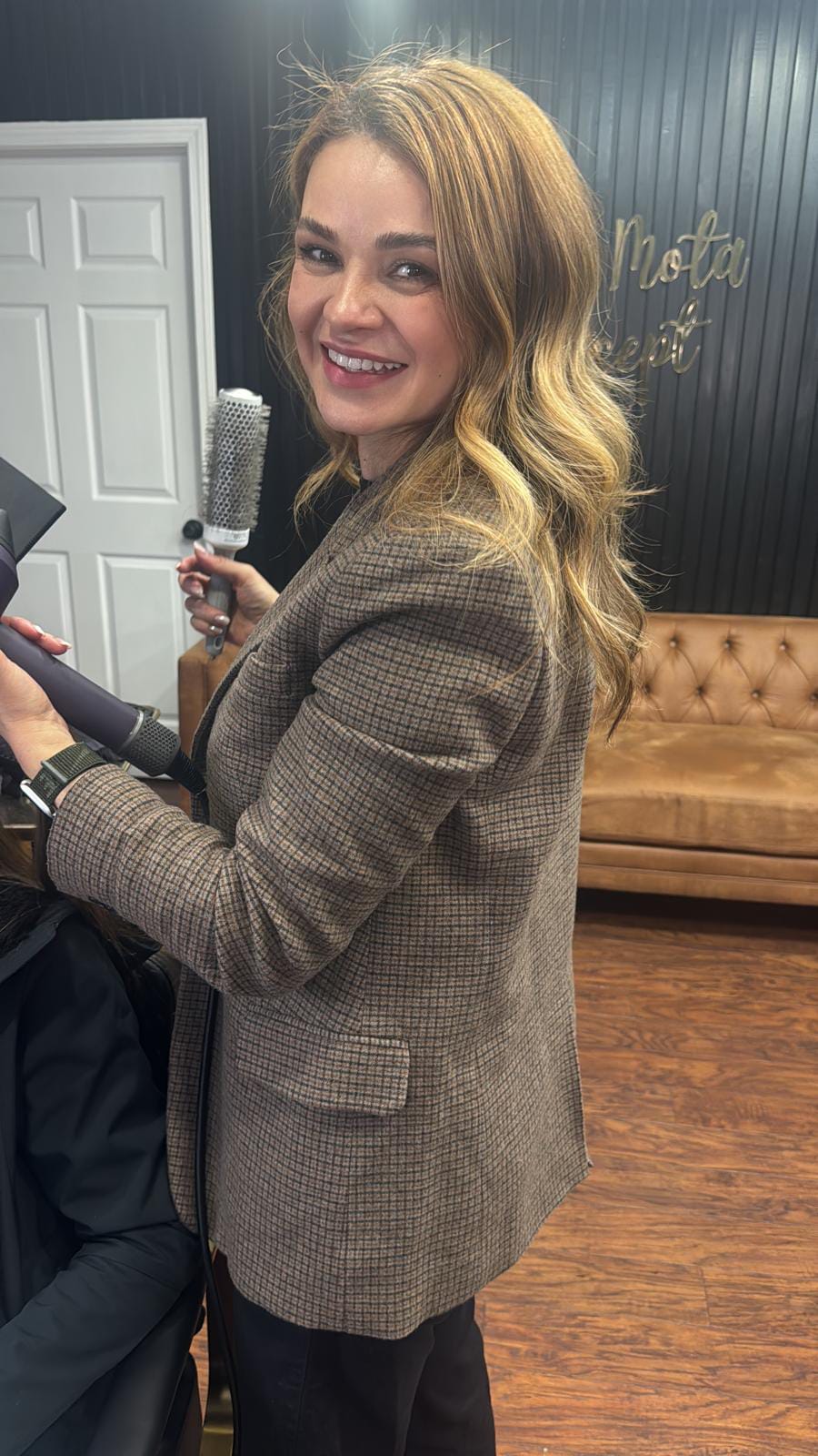 Erika Mota sorrindo de blazer xadrez, com uma escova de cabelo na mão